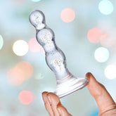 In a hand, the Gläs Triple Play 4" Beaded Glass Butt Plug Dildo features prominently with its transparent tempered glass construction, consisting of three rounded segments and a flat base. It stands out against a blurred backdrop of multicolored bokeh lights, with its slightly sparkly, reflective surface hinting at elegant craftsmanship.
